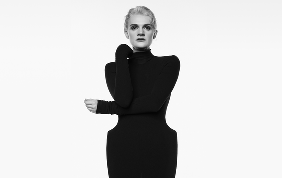A black and white image of a blond woman in sculptural black dress with one folded arm across her body