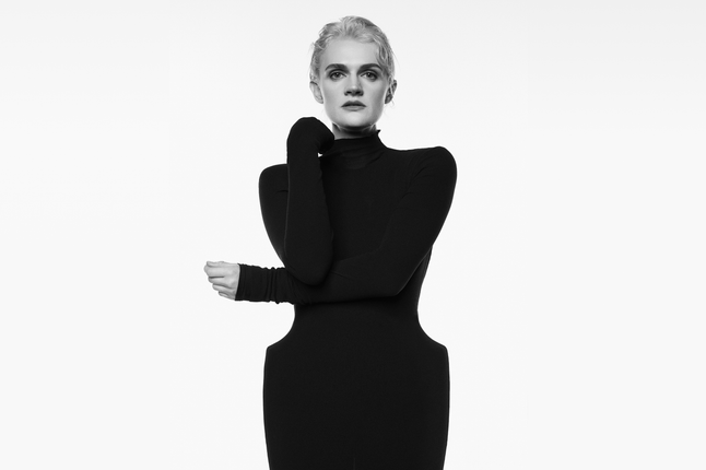 A black and white image of a blond woman in sculptural black dress with one folded arm across her body