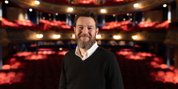 David Greig, a white man with a beard, stands in front of an illuminated auditorium.